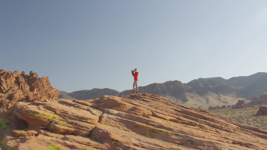 A white woman with blonde hair and an orange sweatshirt with shorts is standing on a mountainside taking photos