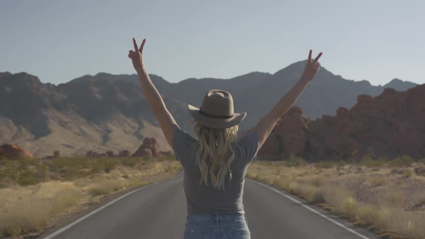 Woman in cowboy hat standing on desert highway holds up both hands in peace signs