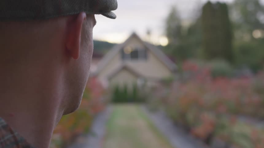 Man with head turned away from the camera watches a house at the end of a garden at sunset