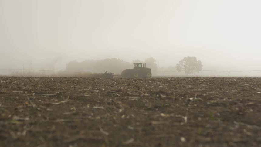 A tractor pulls a plow behind it as it crosses a field on a foggy day