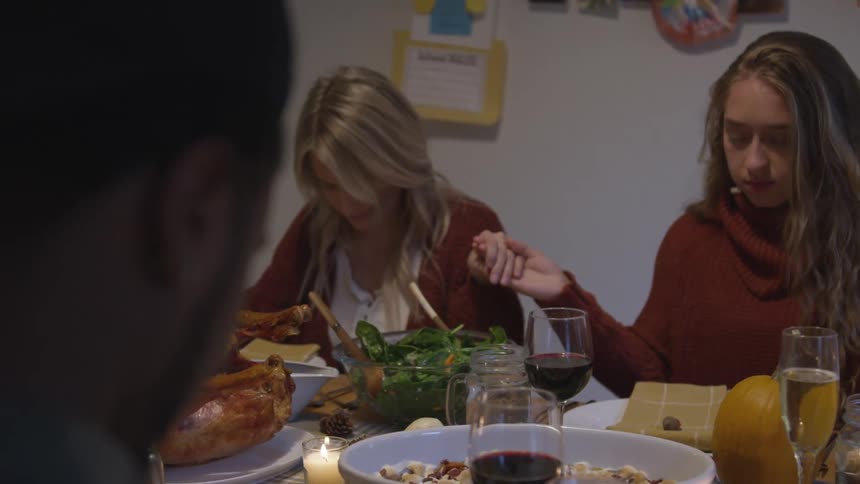 A family holds hand and bows their head in prayer as they sit around a dining table covered in food and wine