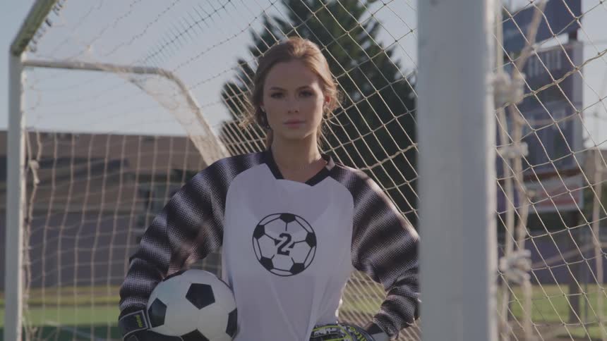 A woman soccer player stands inside a goal holding a ball