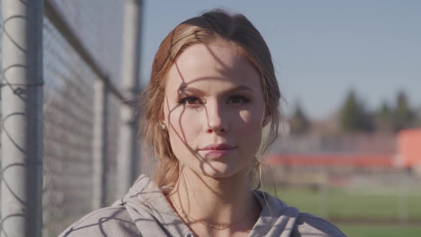 A woman standing and staring next to a fence with trees and the sky in the background