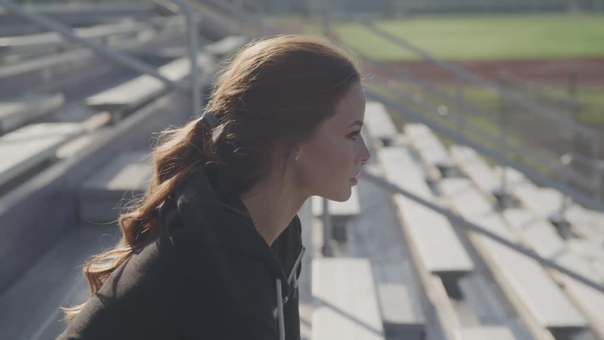 A woman sits alone in the empty bleachers at an outdoor track field