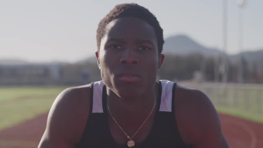 A young black man kneeling on an athletics track in preparation for a sprint raises his head to look into the camera
