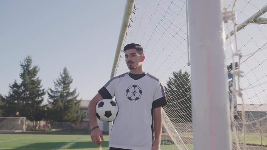 A young hispanic soccer player stands in front of a soccer goal holding a soccer ball as he slowly turns his head to look into the camera