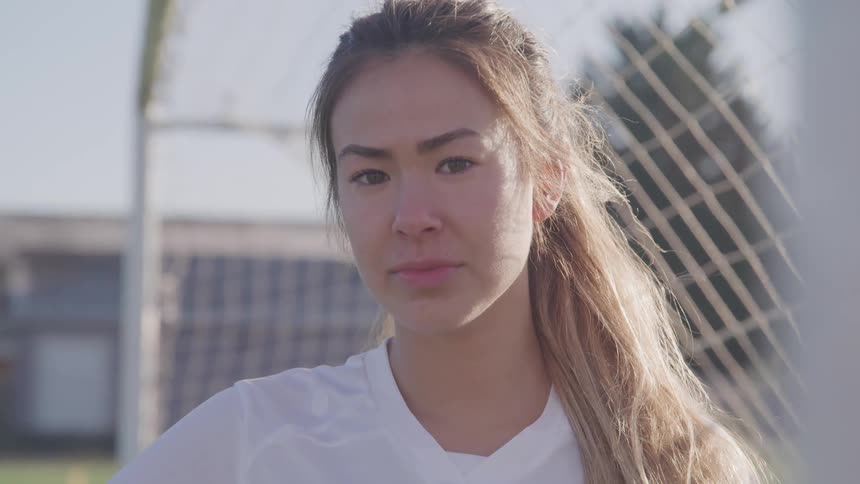 A young asian woman in a jersey looks into the camera while standing in a soccer goal