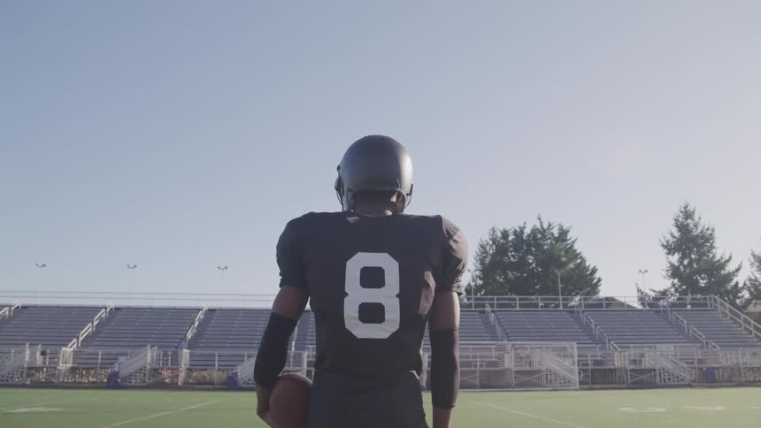 Football player stands in gear holding football and looking out over football field