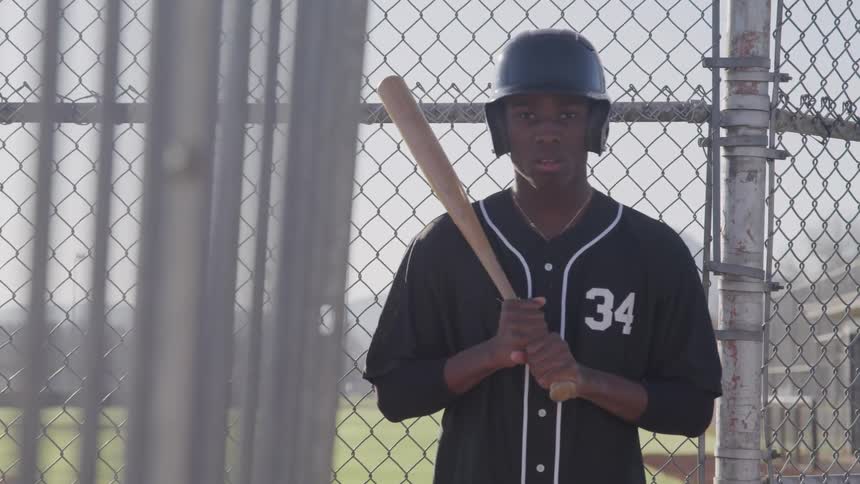 A young black baseball player poses with a baseball bat while looking into the camera