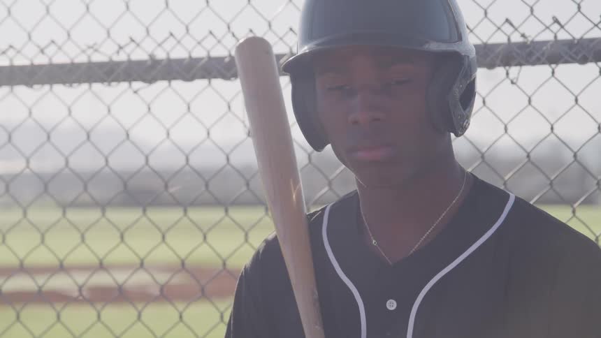 A male baseball player stands by the field fence holding a baseball bat