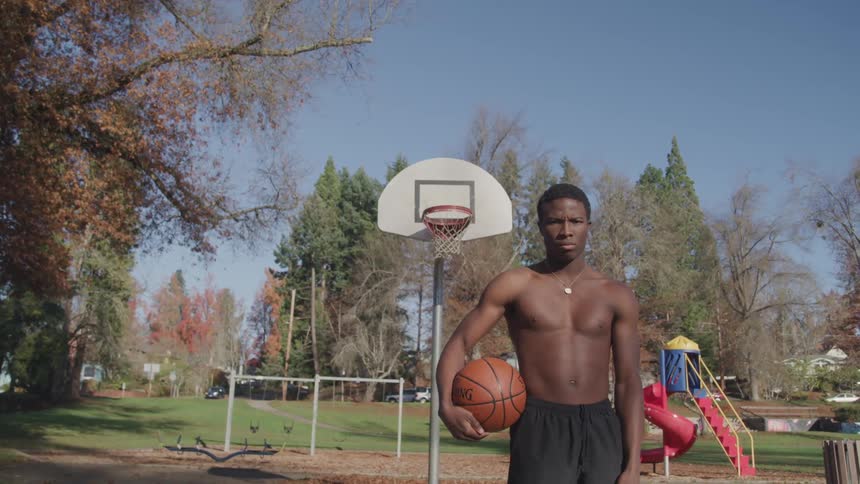 A black teenage boy stands shirtless on a basketball court with a basketball under his arm looking ahead