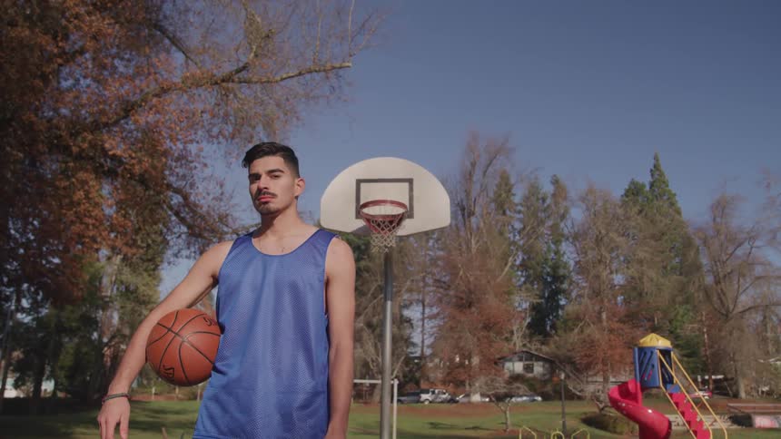 A man poses in front of a basketball hoop at a playground with a basketball under his arm
