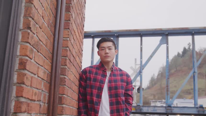 Asian man with a plaid shirt leans against a brick wall and looks at the camera at the train station