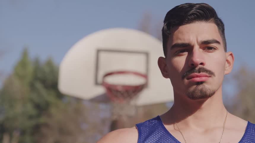 Man with a goatee beard and a blue basketball jersey faces the camera with eye contact in a local basketball court