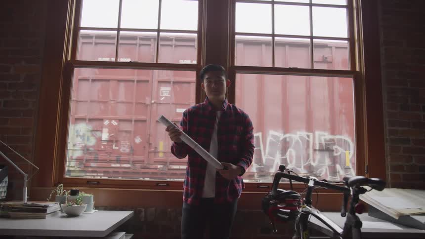 A young asian man holding rolled up paper stands in a front of a window as a train covered in graffiti passes by on the other side