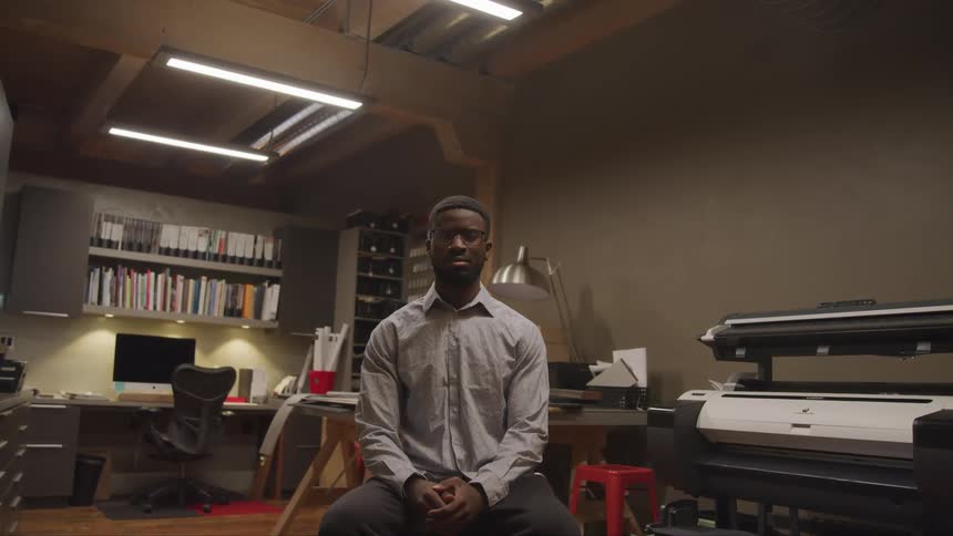 A young male sits in a office workspace on a stool while posing