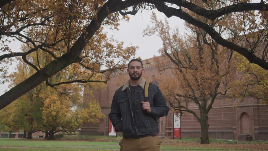 Man with a denim jacket a backpack and a beard stands under an autumn tree on green grass