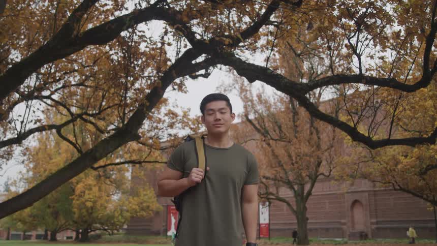 Asian man with a t shirt and a backpack over shoulder looks at the camera smilingly under an autumn tree
