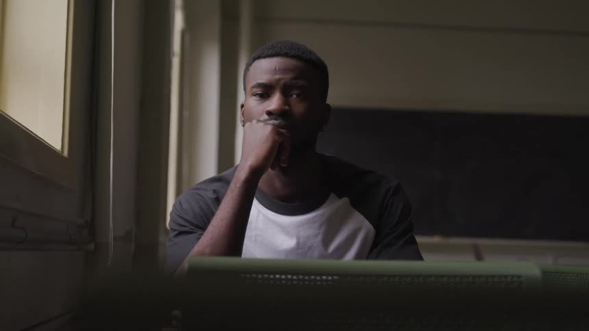 A black male with thick short black hair sits with his closed hand resting on his chin posing in a dim classroom
