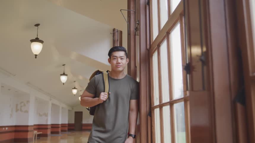 Asian man with a grey t shirt and a backpack leans against windows and faces the camera with eye contact in the corridor