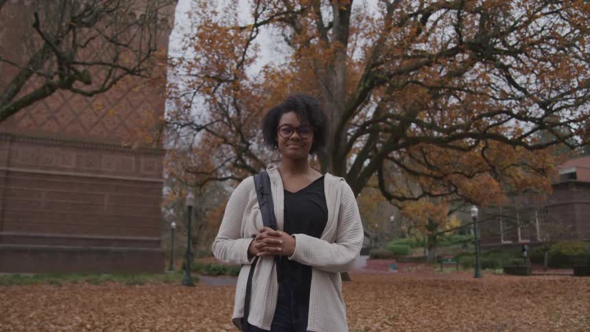 A black woman is standing in the middle of campus with leaves fallen around her