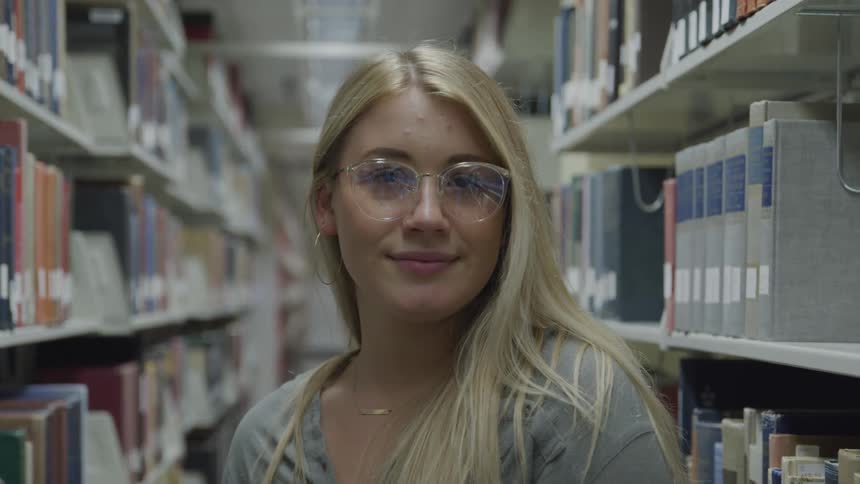Blonde woman with glasses faces the camera with eye contact in the library