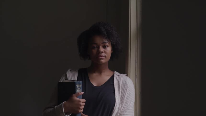 Black woman with curly hair holds a book and looks at the camera in dim lit classroom