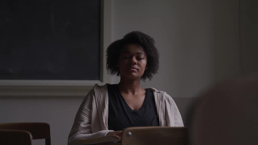 Black woman with short curly hair sits in a chair and looks at the camera in a dim lit classroom
