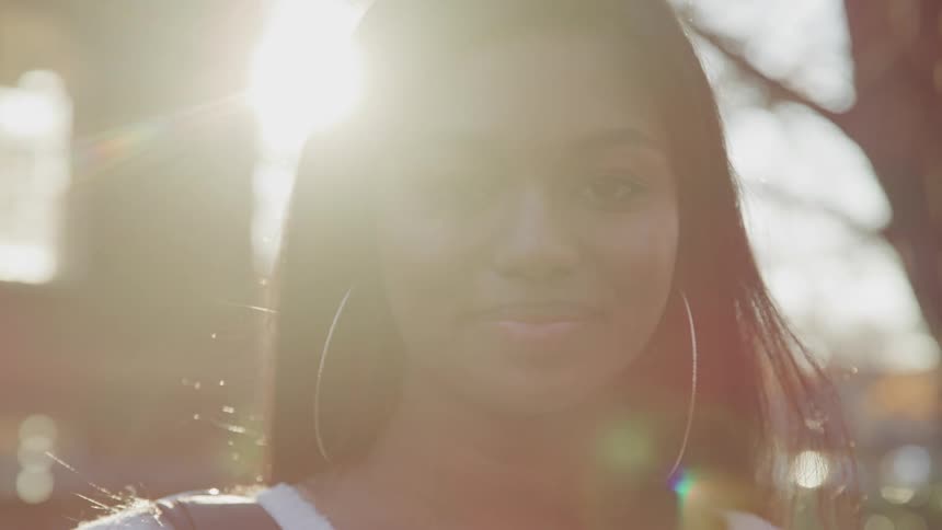 Dark skinned woman with hoop earrings faces the camera with eye contact at sunset