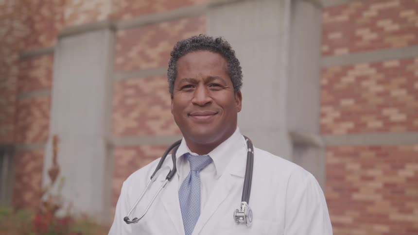 Dark skinned doctor with a white lab coat and a stethoscope looks at the camera warmly in front of the hospital