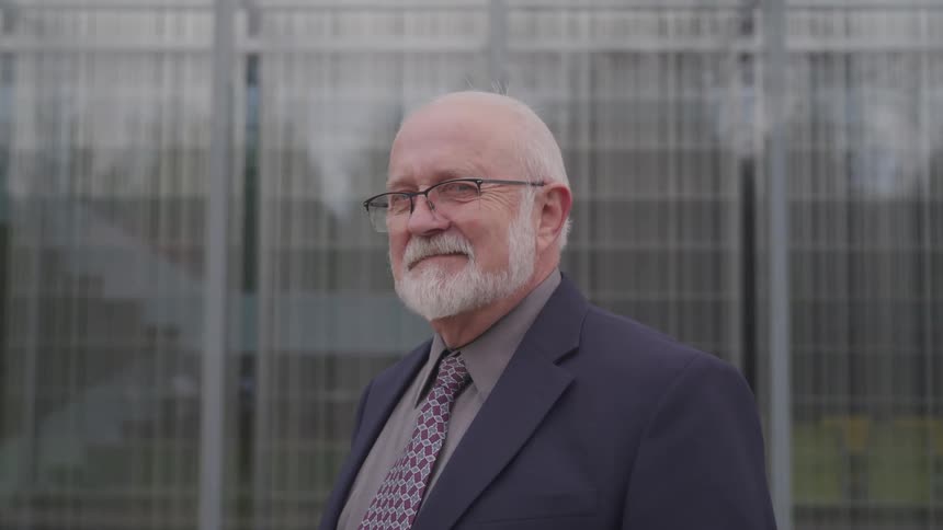 Man with white beard and mustache stands in front of building windows wearing suit and necktie