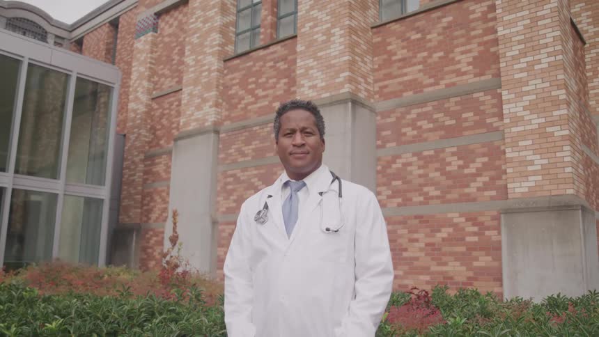 Middle aged doctor with stethoscope and a white lab coat stands and looks at the camera in front of the hospital