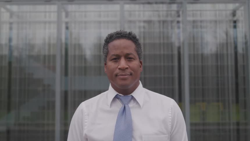 Dark skinned man with white suit and a blue tie faces the camera with eye contact