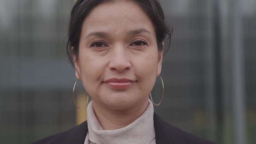 Woman with a turtleneck sweater a blazer and hoop earrings faces the camera with eye contact