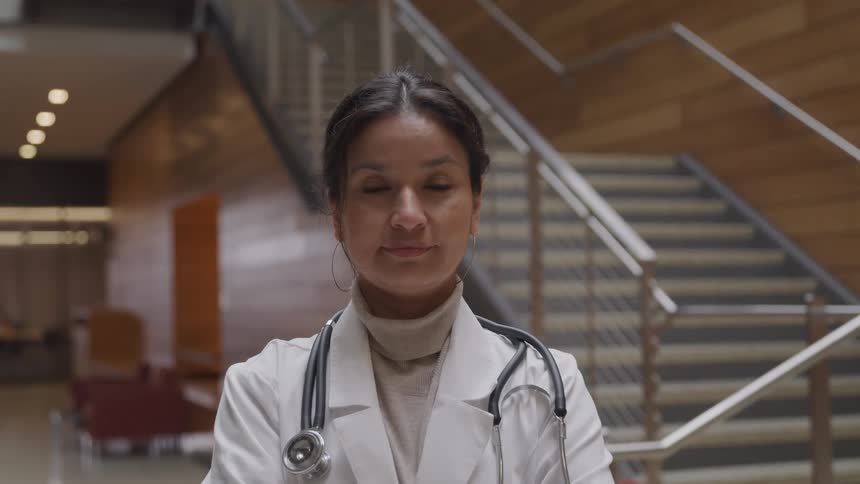 Doctor wearing a lab coat and stethoscope stands in an hospital hall looking at the camera