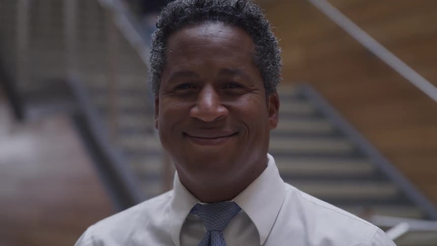 A doctor stands at the bottom of a set of stairs and smiles as a person walks down the stairs