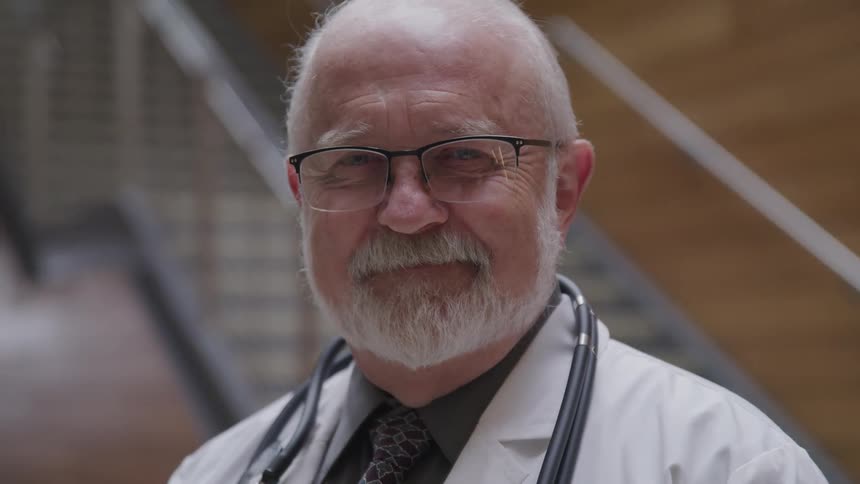 An elderly doctor stands at the bottom of a set of stairs and smiles