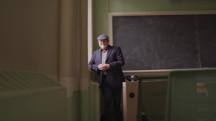 An older man stands at the front of a classroom next to a window