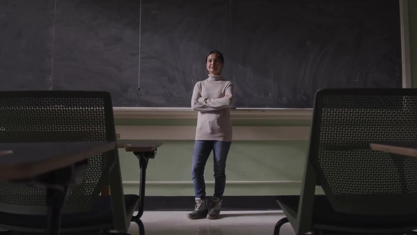 Woman with a grey turtleneck sweater jeans stands arms crossed in front of the blackboard in the classroom