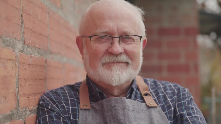 An older man with a beard wearing an apron leans against a brick wall while posing