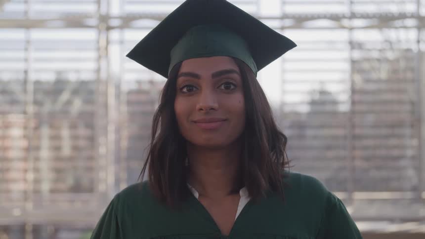A college graduate wears her green cap and gown while posing on campus in front of a large window