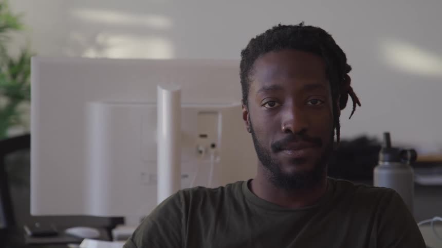 Black man with braided hair and a beard faces the camera with eye contact with a computer monitor in the background