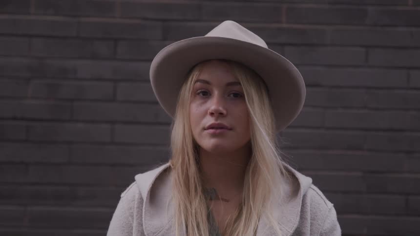 Blonde woman wearing hat standing in front of brick wall turns to face forward