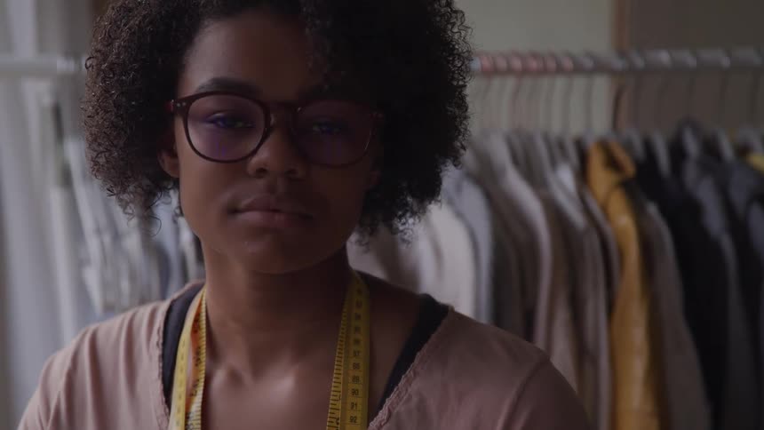 A young black woman wearing glasses poses for the camera in front of some clothing