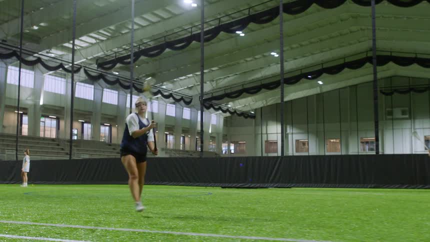 A group of teenage girls play lacrosse on an indoor field
