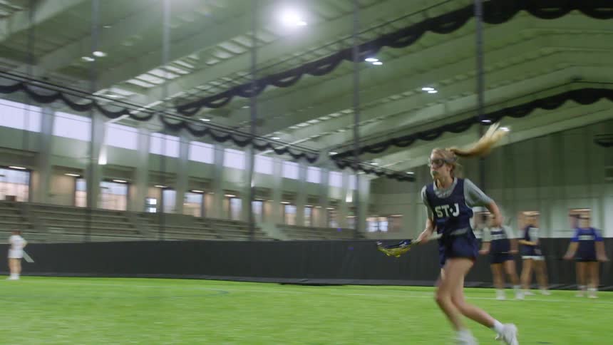 Young women do lacrosse drills on an indoors field during the day