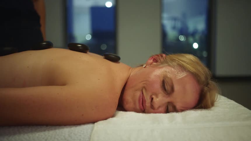 A woman smiles as a masseuse places stones along her spine
