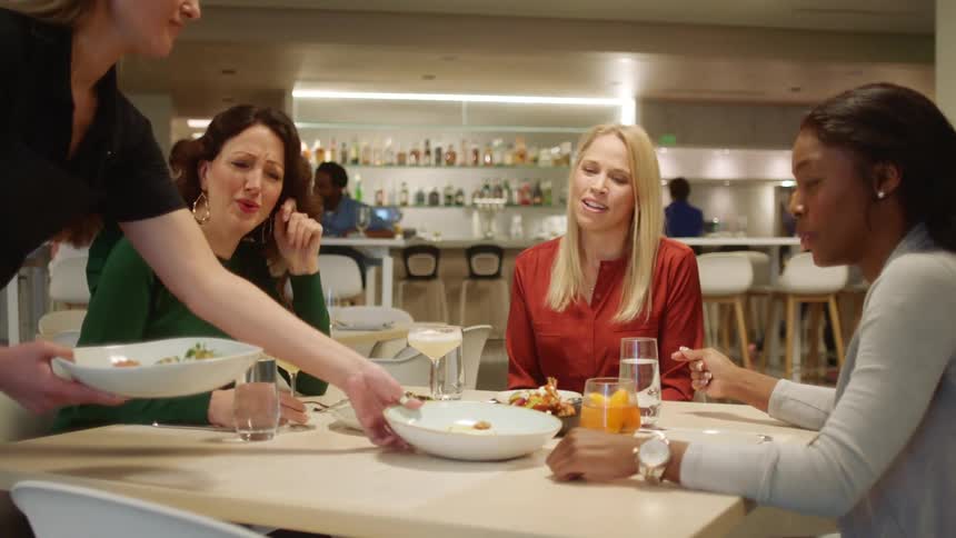 Three female friends laugh and talk while sitting at a table in a restaurant then thank the waitress as she sets down two plates of appetizers on the table