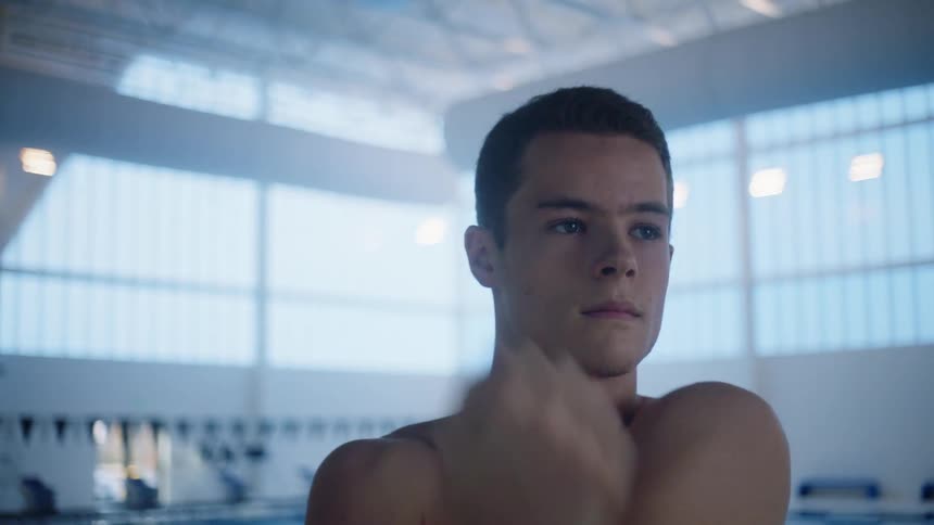 A shirtless young man does stretches while standing at the side of a pool in an indoor pool