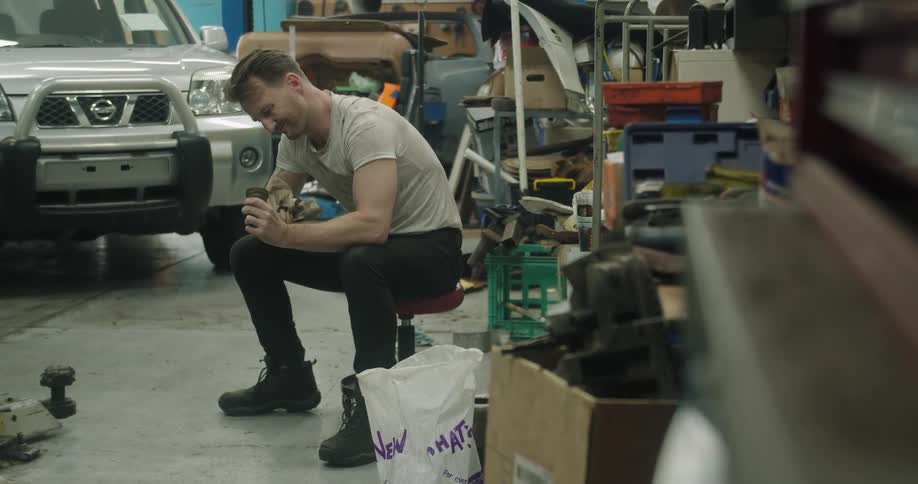 A mechanic sits on a stool near a silver sport utility vehicle and uses a rag to wipe an auto part clean